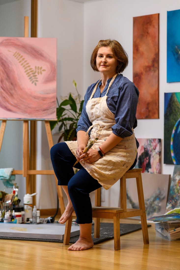 Portrait of Mona Barbu in her studio, seated on a wooden stool with paintbrushes in hand